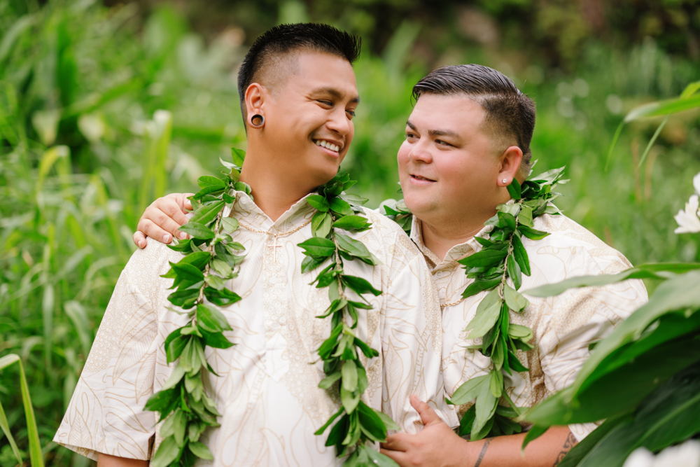 Tylor and Austin smiling in white aloha shirts and green maile lei among tropical plants